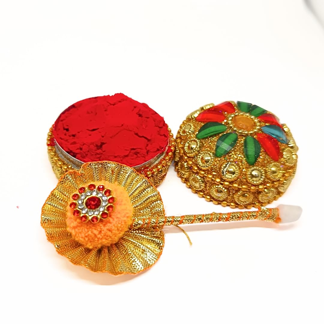 A traditional Indian decorative set featuring a small round gold-toned pot filled with bright red powder, a matching ornate lid with gold detailing and red and green floral accents, and a decorative gold-toned tool with a fluffy orange pom-pom and red jewel center.
