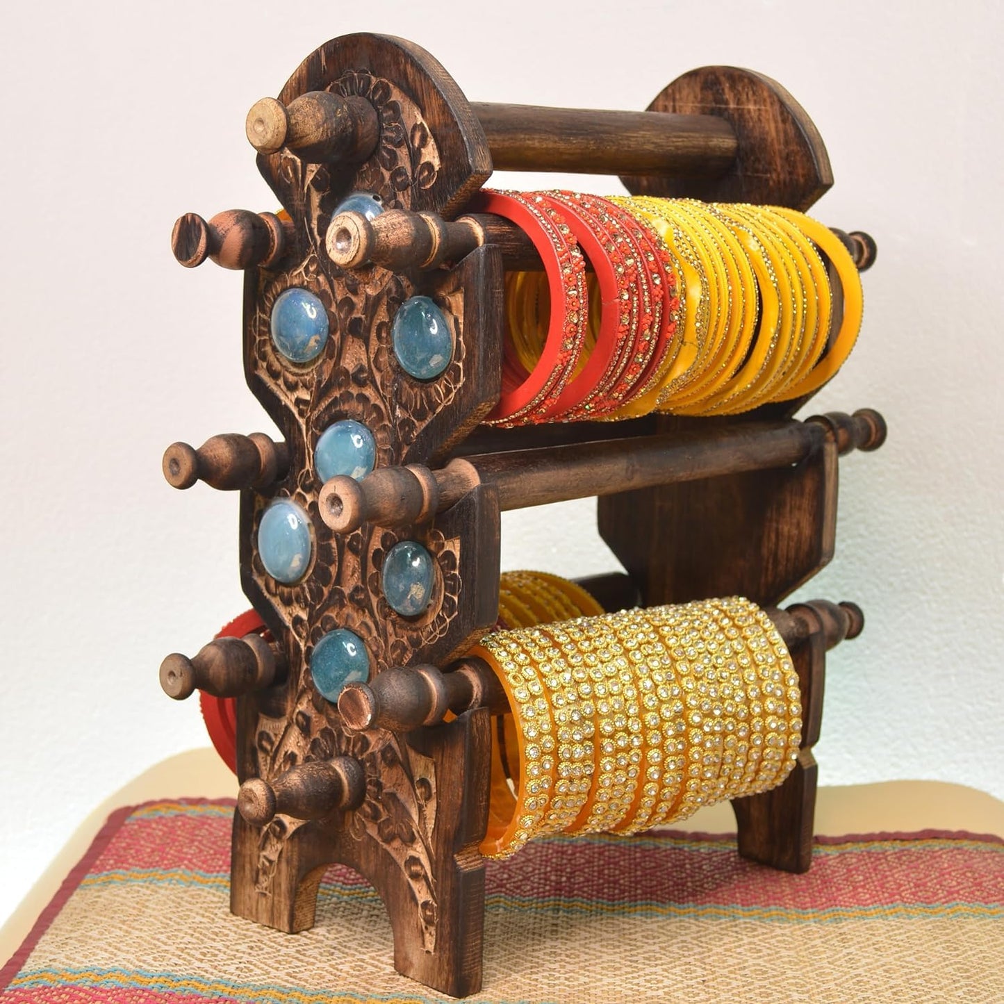 An ornate dark brown carved wooden bangle rack with carved details and blue stone accents, holding multiple stacks of colorful bangles on its three tiers, set against a plain light background.