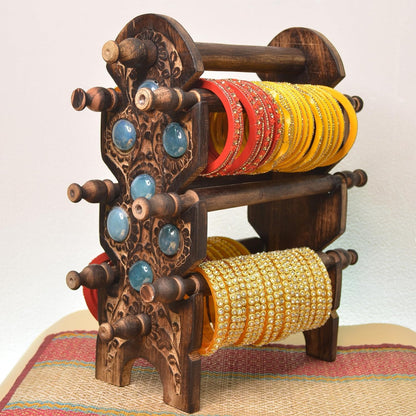 An ornate dark brown carved wooden bangle rack with carved details and blue stone accents, holding multiple stacks of colorful bangles on its three tiers, set against a plain light background.