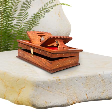 A small carved wooden box with an open hinged lid, brass latch, and brass corner accents, placed on a light stone slab with a blurred white vase and green fern in the background.