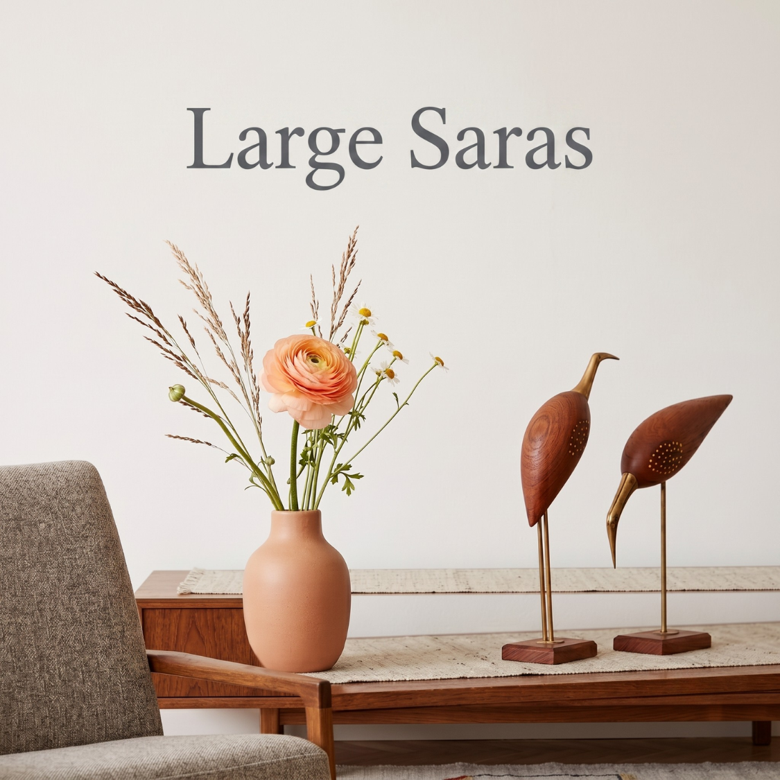 A styled interior scene with a terracotta vase holding dried grasses and a peach flower on a wooden table, next to two wooden bird sculptures on brass stands. The words 'Large Saras' appear above on a light wall.