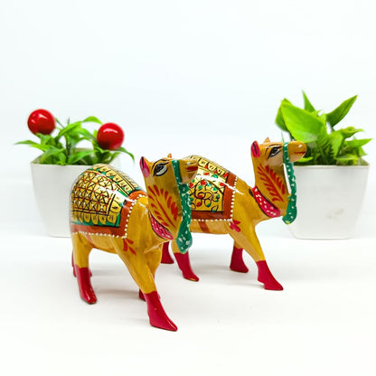Two hand-painted camel figurines decorated with traditional Indian patterns in yellow, red, green, and orange, set against a white background with decorative potted plants.